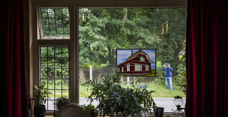 Een raam met deels glas-in-lood, plantjes op de vensterbank en buiten op straat een persoon die het huis fotografeert