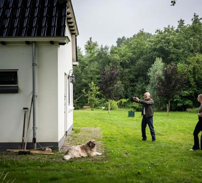 Drie personen en een hond staan in een grote tuin, waarbij een persoon zijn camera richt om een foto te maken van een gebouw.