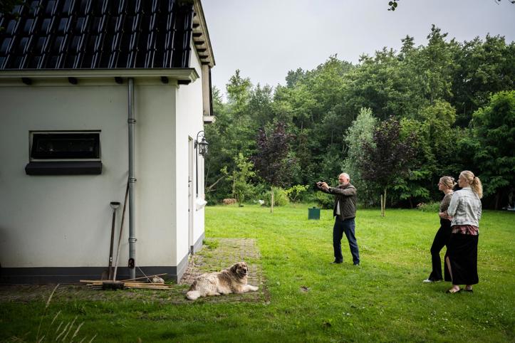 Drie personen en een hond staan in een grote tuin, waarbij een persoon zijn camera richt om een foto te maken van een gebouw.