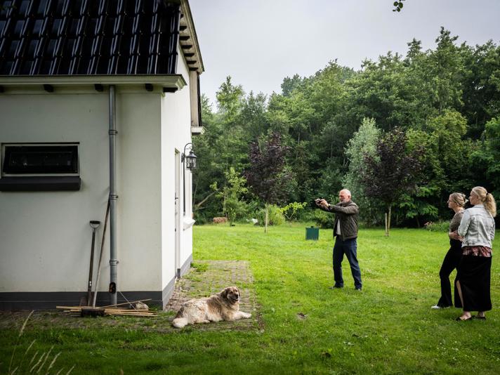 Drie personen en een hond staan in een grote tuin, waarbij een persoon zijn camera richt om een foto te maken van een gebouw.