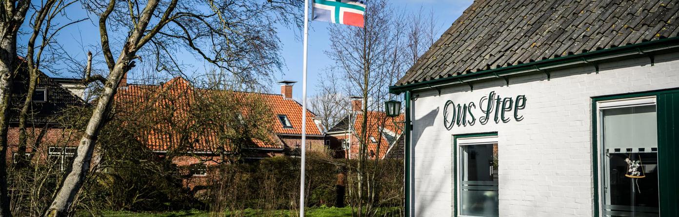 Een wapperende Groningse vlag op stok in de tuin van een wit huis met daarop de tekst: Ons Stee.