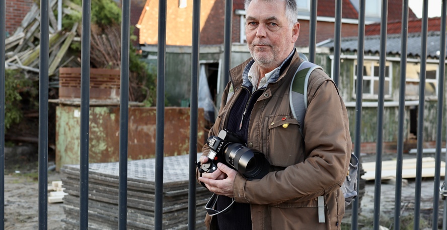 Jan Zeeman staat met zijn fotocamera in de hand voor een hek om een bouwplaats