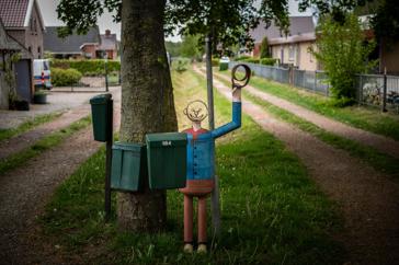 Een kunstwerk van een vrolijke man met zwaaiende arm bij drie brievenbussen langs de weg