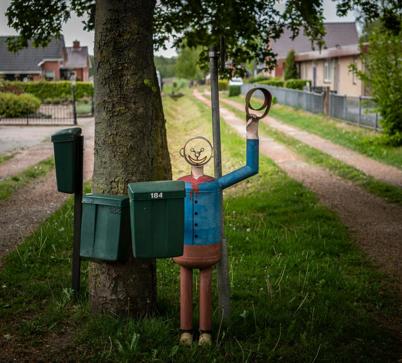 Een kunstwerk van een vrolijke man met zwaaiende arm bij drie brievenbussen langs de weg