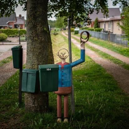 Een kunstwerk van een vrolijke man met zwaaiende arm bij drie brievenbussen langs de weg