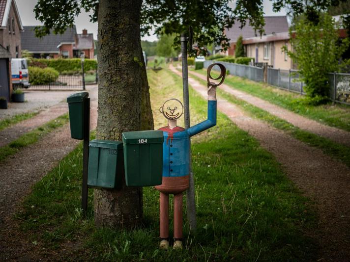 Een kunstwerk van een vrolijke man met zwaaiende arm bij drie brievenbussen langs de weg