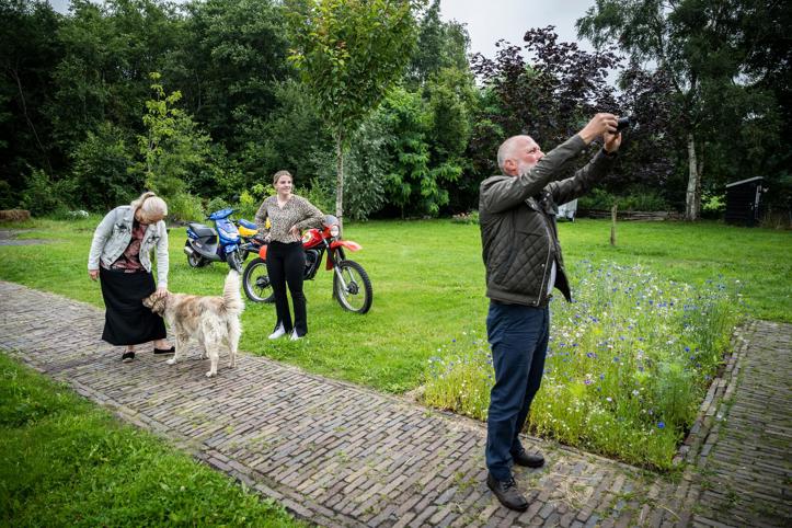 Drie personen en een hond staan in een grote tuin, waarbij een persoon zijn camera richt om een foto te maken.