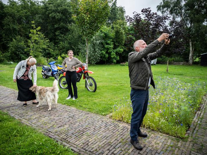 Drie personen en een hond staan in een grote tuin, waarbij een persoon zijn camera richt om een foto te maken.