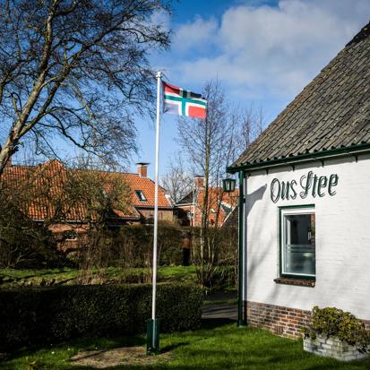 Een wapperende Groningse vlag op stok in de tuin van een wit huis met daarop de tekst: Ons Stee.