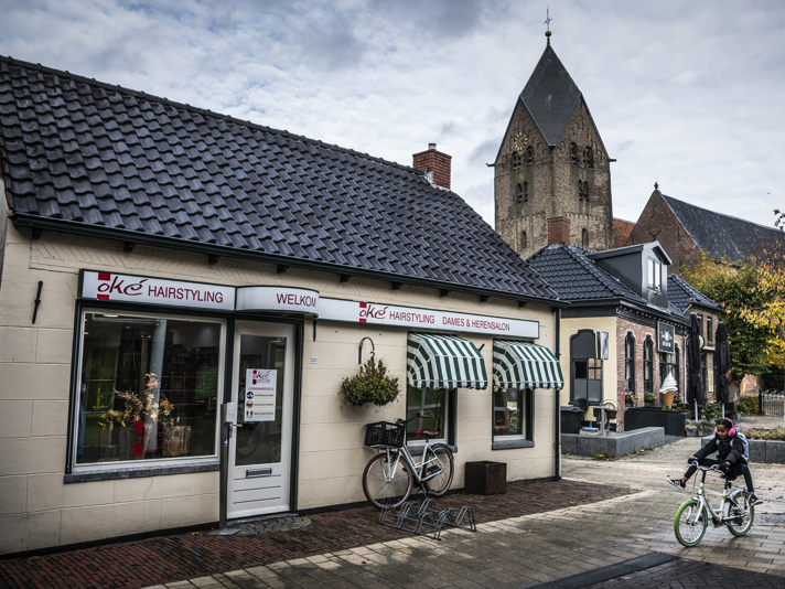 Een dorpsplein met een kapsalon, een kind op de fiets en een kerktoren die boven de gebouwen uitsteekt.