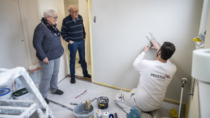 Twee personen kijken lachend naar een meneer die stucwerkzaamheden uitvoert in een kamer.