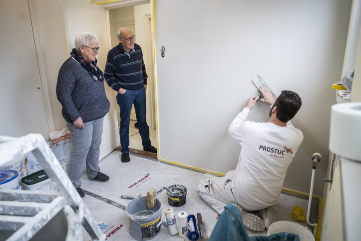 Twee personen kijken lachend naar een meneer die stucwerkzaamheden uitvoert in een kamer.