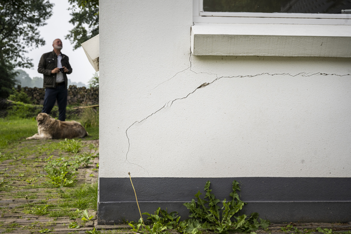 Grote scheur aan de buitenwand van een huis waarnaast een persoon staat en een hond op de grond eronder ligt. 